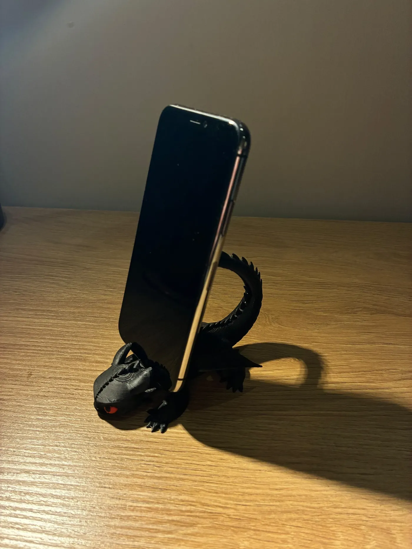 Black 3D printed dragon phone stand with red eyes holding a smartphone on a wooden desk