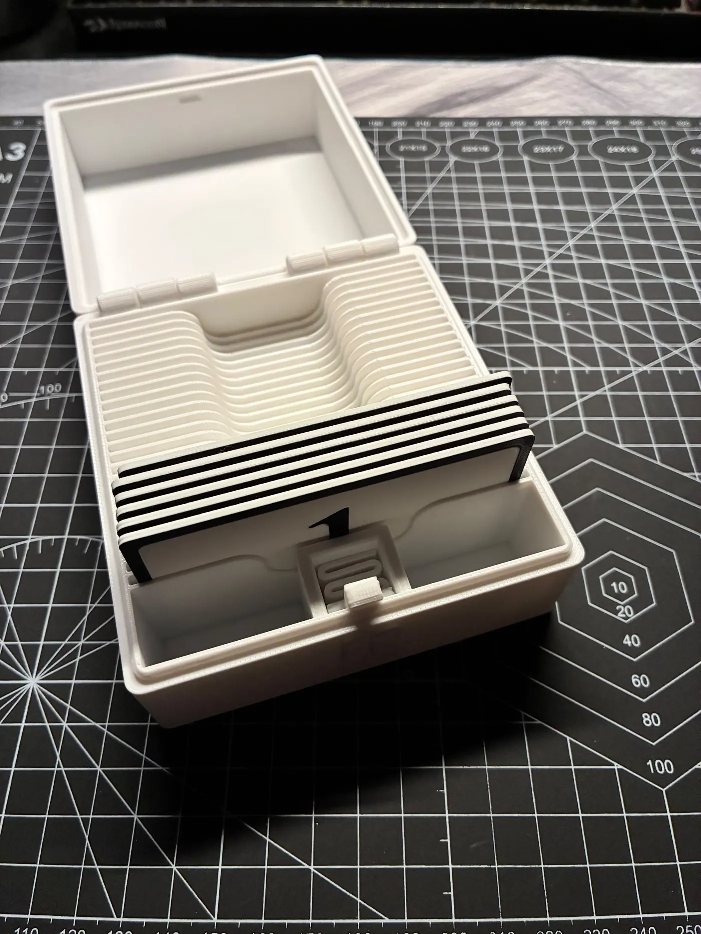 Open white 3D printed box with hinged lid, curved card rack and stack of black-edged number cards