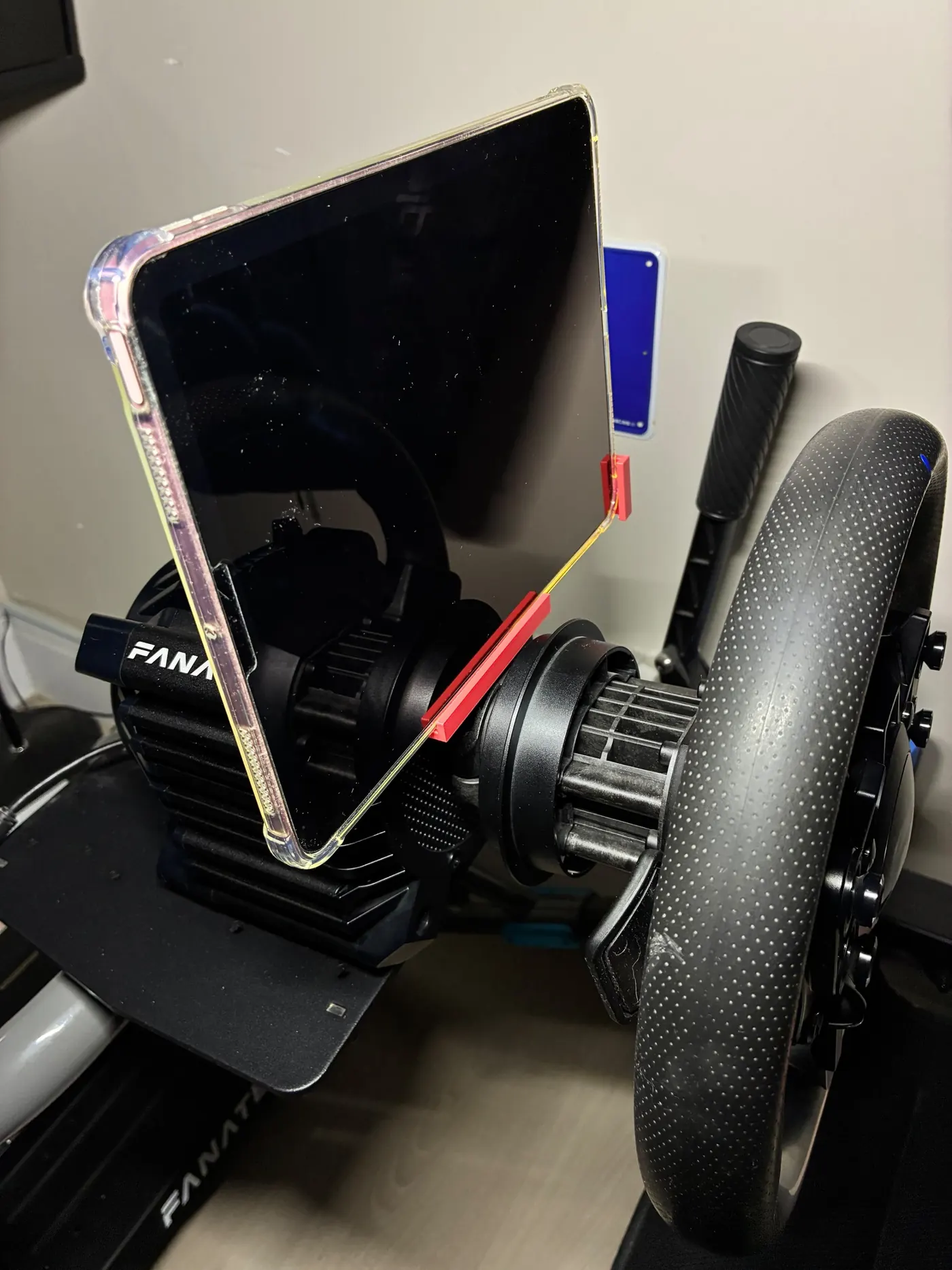 Tablet in a clear case held by red 3D printed clips on a sim-racing wheelbase, steering wheel in front