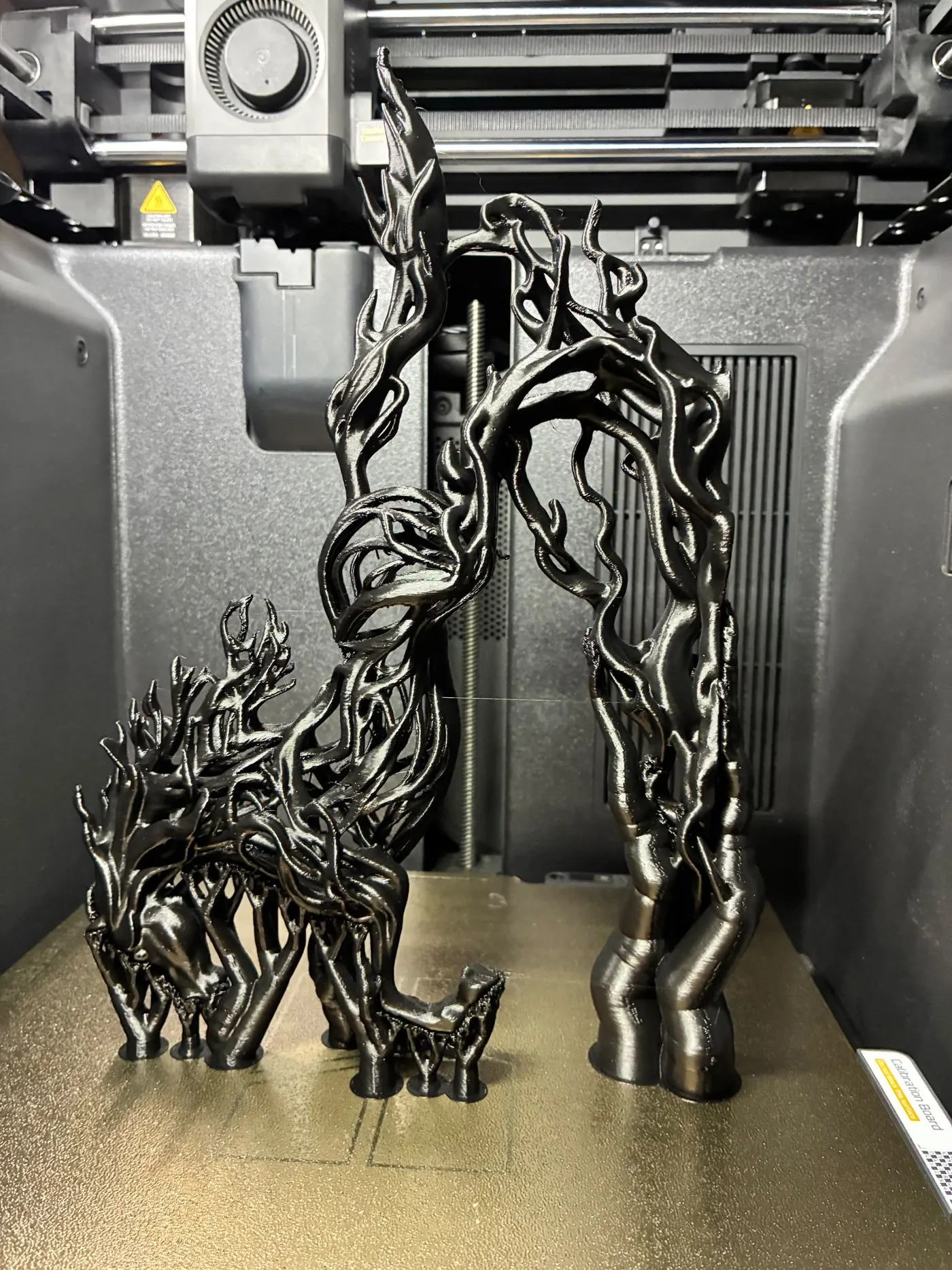 Black 3D printed sculpture of a stag merging into branching roots and an arched tree on the print bed